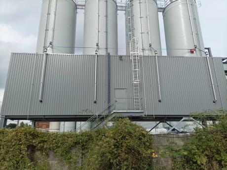 Four white industrial silos lined up on a metal platform with a safety ladder and pipes, in front of a grey panel façade.